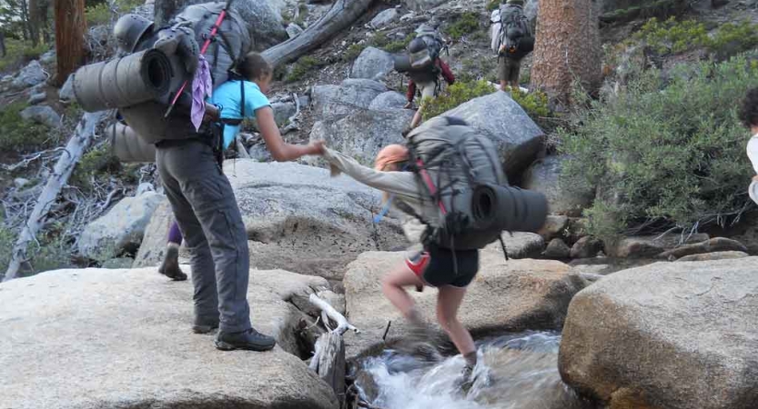 One backpacker helps another cross a rocky stream.